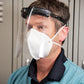 Picture of person wearing a face shield over a white mask; person is wearing blue scrubs and grey lockers in the background